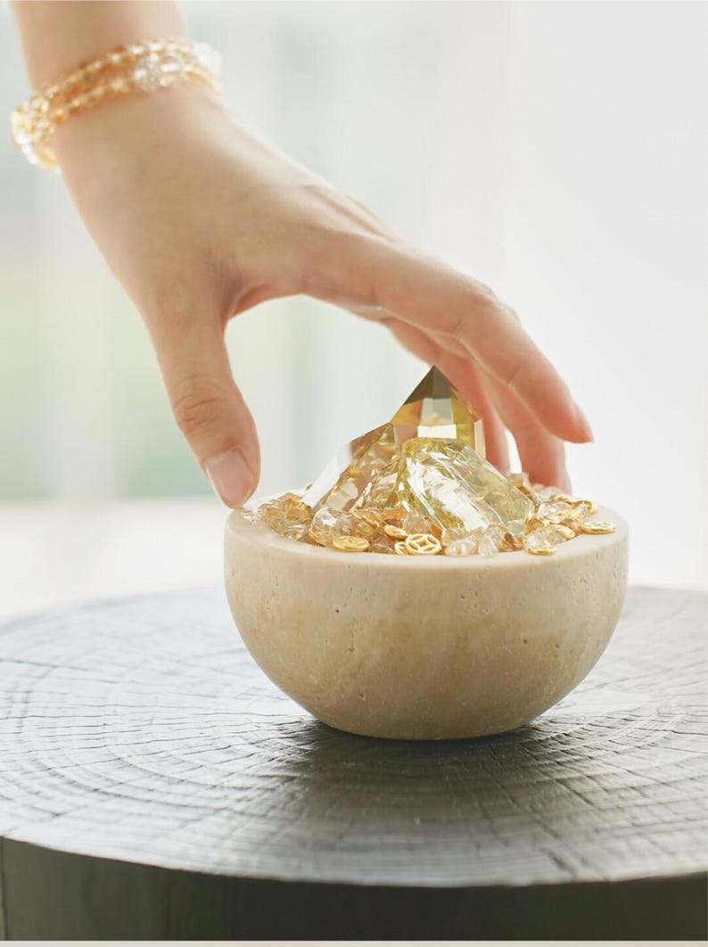 May include: A beige, semi-spherical bowl filled with golden crystals and small, decorative gold pieces. A large, faceted crystal sits in the center. A hand hovers over the bowl, and a beaded bracelet is visible in the background.