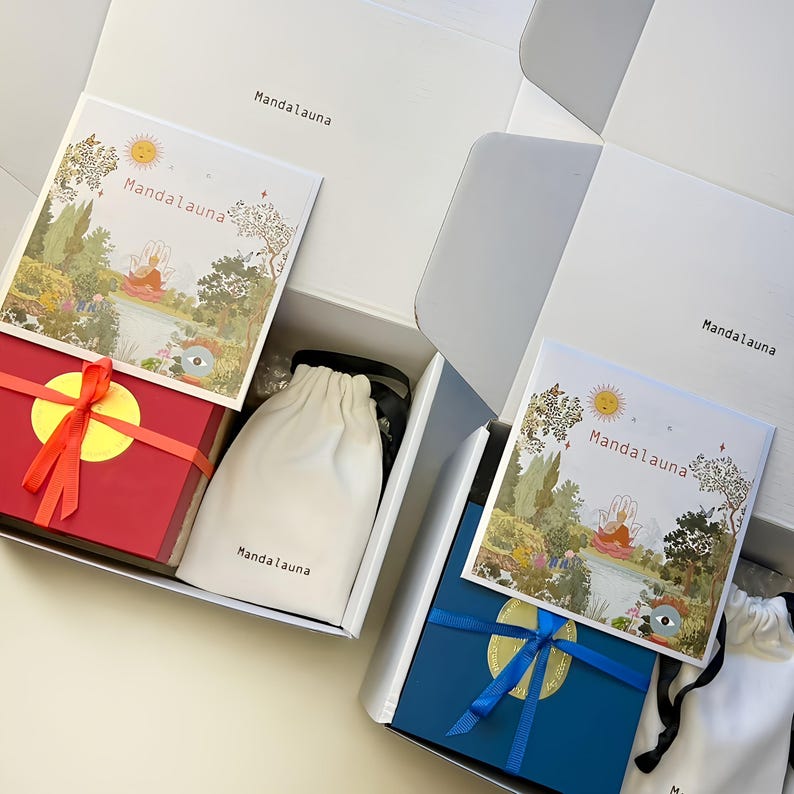 May include: Two open white boxes with gifts inside. One box contains a red gift box tied with an orange ribbon and a white drawstring bag. The other box has a blue gift box with a blue ribbon. Each box has a card with the text "Mandalauna".