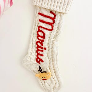 May include: Cream cable knit Christmas stocking with the name "Marius" embroidered in red. A small reindeer head with antlers and a red nose is attached near the toe. The stocking has a folded-over cuff.