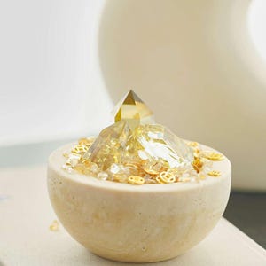 May include: A decorative, beige, half-sphere bowl filled with golden crystals and small, gold-colored, circular embellishments. A clear, pyramid-shaped crystal sits atop the crystals. The bowl is on a neutral-colored surface.