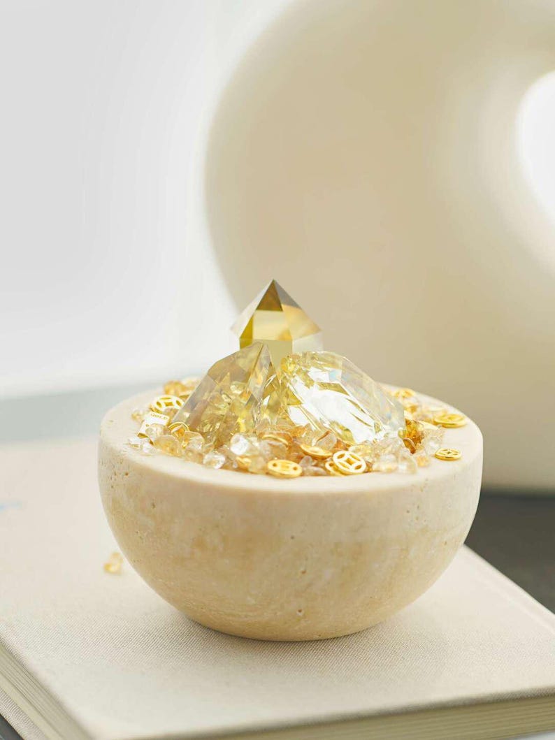 May include: A decorative, beige, half-sphere bowl filled with golden crystals and small, gold-colored, circular embellishments. A clear, pyramid-shaped crystal sits atop the crystals. The bowl is on a neutral-colored surface.