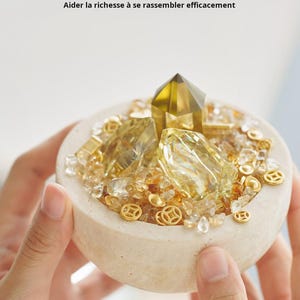 May include: A small, beige bowl filled with golden crystals, small gold coins, and a large, faceted yellow crystal. The bowl is held by hands. Text in French reads: "Aider la richesse &agrave; se rassembler efficacement."