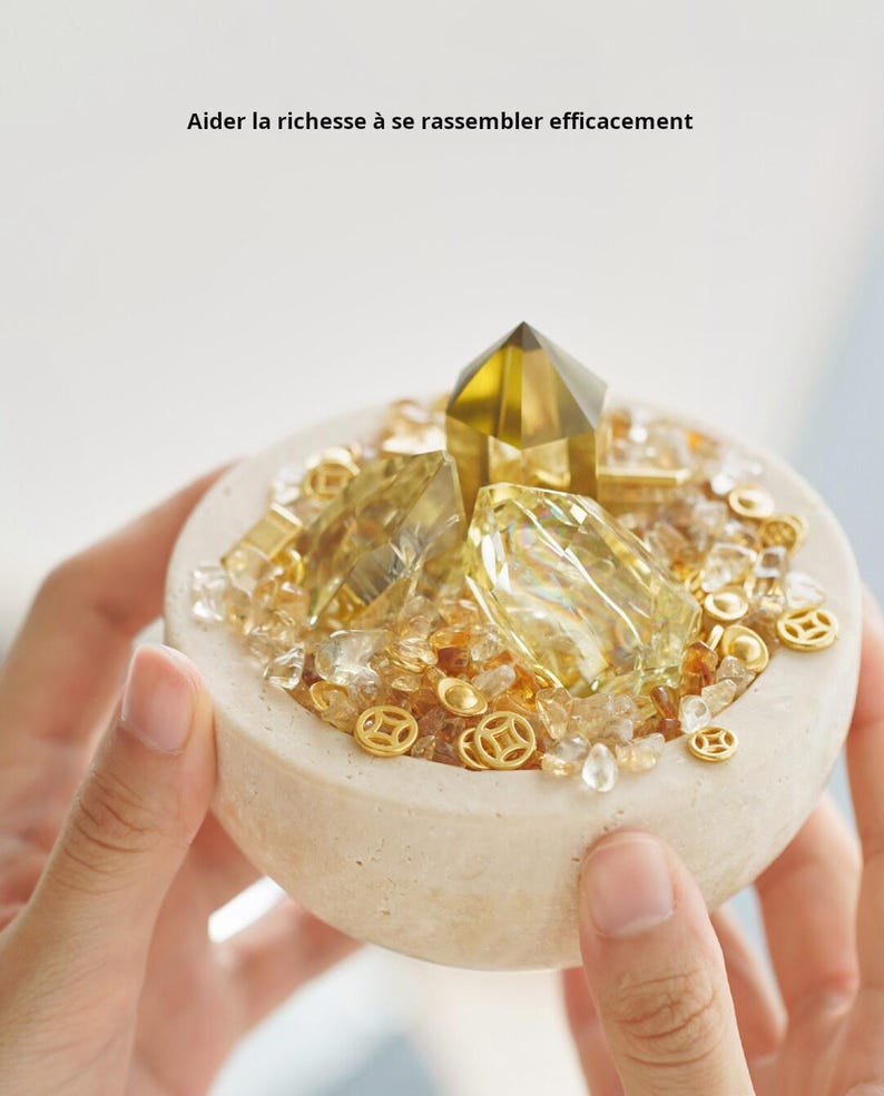 May include: A small, beige bowl filled with golden crystals, small gold coins, and a large, faceted yellow crystal. The bowl is held by hands. Text in French reads: "Aider la richesse &agrave; se rassembler efficacement."