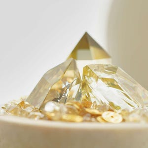 May include: Close-up of a decorative arrangement featuring clear, faceted crystal pyramids and small, golden-colored stones. The crystals are arranged on a light-colored, cylindrical base, creating a layered effect. The overall aesthetic is minimalist and elegant.