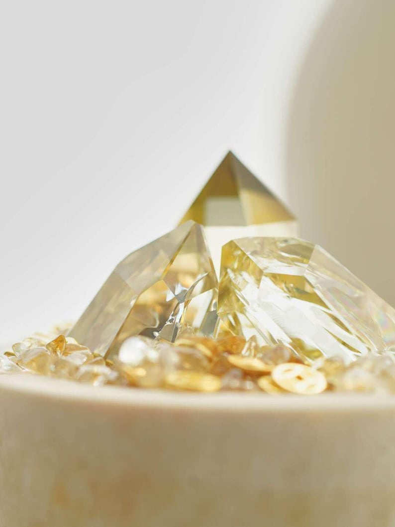 May include: Close-up of a decorative arrangement featuring clear, faceted crystal pyramids and small, golden-colored stones. The crystals are arranged on a light-colored, cylindrical base, creating a layered effect. The overall aesthetic is minimalist and elegant.