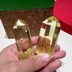 May include: Two yellow-toned crystal points are held in a hand. The crystals have a faceted, geometric shape. A green box and a red box are in the background. The crystals are a light, translucent yellow.