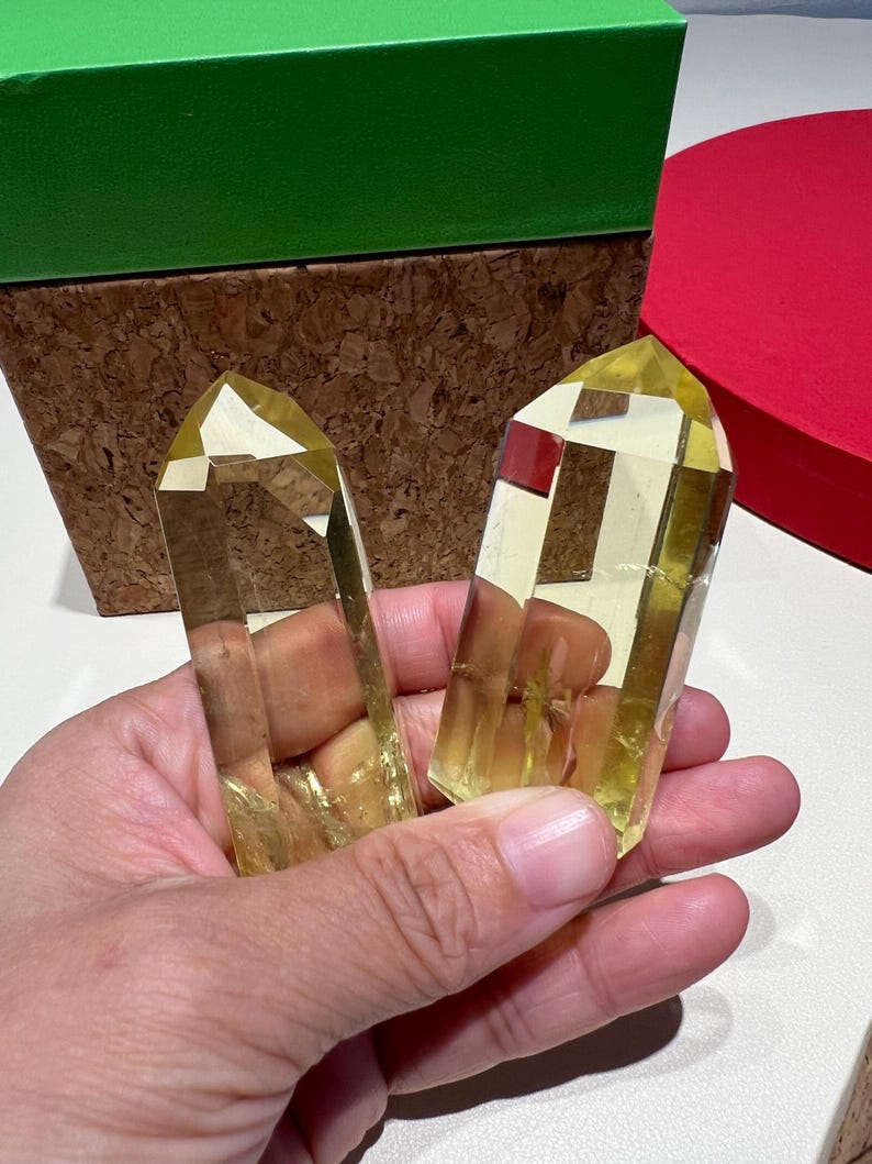 May include: Two yellow-toned crystal points are held in a hand. The crystals have a faceted, geometric shape. A green box and a red box are in the background. The crystals are a light, translucent yellow.