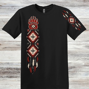 May include: Black t-shirt with Native American-inspired designs. Features a bear paw print, geometric patterns in red, white, and black, and feather accents. The design is on the front and sleeve of the shirt.