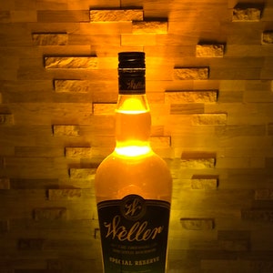 May include: A lit-up Weller Special Reserve bourbon bottle. The bottle has a black and gold label with the brand name and text. The bottle is illuminated from within, casting a warm glow against a textured wall.