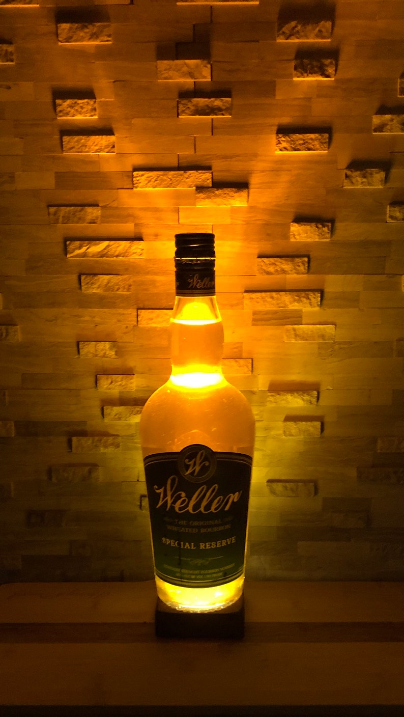 May include: A lit-up Weller Special Reserve bourbon bottle. The bottle has a black and gold label with the brand name and text. The bottle is illuminated from within, casting a warm glow against a textured wall.