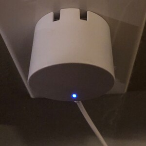 May include: A white, cylindrical device plugged into a wall socket. The device has a rounded base and a notched top. A thin, white cable extends from the base, and a small blue light is illuminated. The device appears to be a charger or adapter.