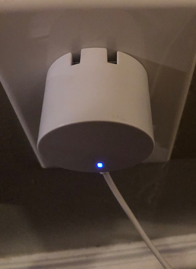 May include: A white, cylindrical device plugged into a wall socket. The device has a rounded base and a notched top. A thin, white cable extends from the base, and a small blue light is illuminated. The device appears to be a charger or adapter.