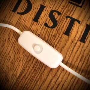 May include: A white inline switch with a rounded button, attached to a white cable. The switch is set against a wooden surface with the word "DISTILL" in black lettering.