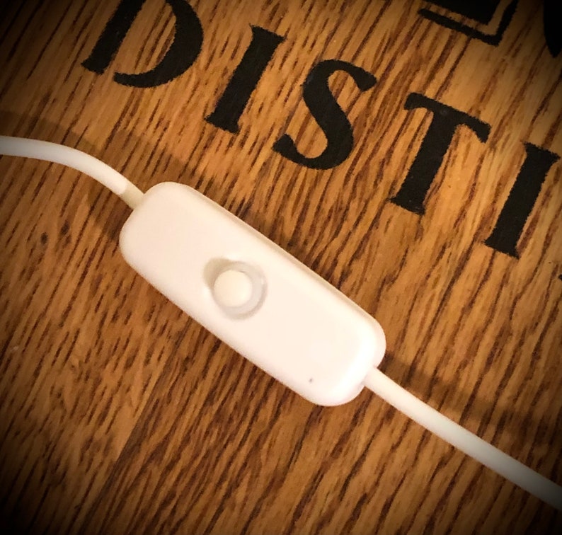 May include: A white inline switch with a rounded button, attached to a white cable. The switch is set against a wooden surface with the word "DISTILL" in black lettering.