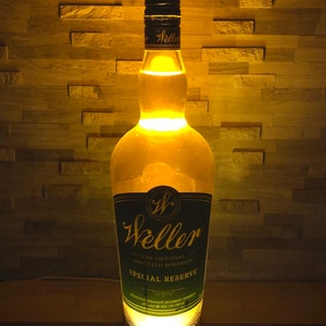 May include: A lit Weller Special Reserve bourbon bottle lamp. The clear bottle has a black and green label. The lamp gives off a warm, yellow glow, lighting up the bottle's contents and the surrounding area. The lamp is on a black base.