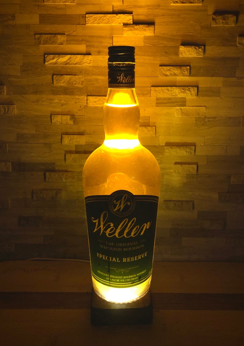 May include: A lit Weller Special Reserve bourbon bottle lamp. The clear bottle has a black and green label. The lamp gives off a warm, yellow glow, lighting up the bottle's contents and the surrounding area. The lamp is on a black base.