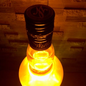 May include: A clear glass bottle with a black cap, lit from within with a warm yellow glow. The cap has a gold emblem and the word "Weller". The bottle is set against a textured, brick-like background.