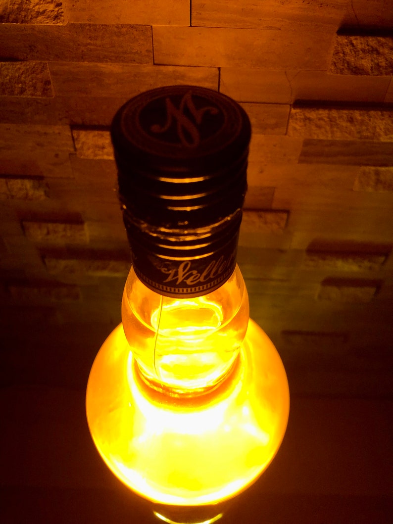 May include: A clear glass bottle with a black cap, lit from within with a warm yellow glow. The cap has a gold emblem and the word "Weller". The bottle is set against a textured, brick-like background.