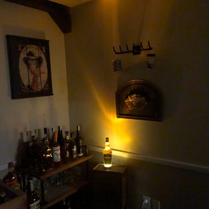 May include: A room with a drinks cabinet and a bottle of liquor lit up on a small wooden stand. A vintage painting and a decorative wall hanging are also visible. The room is dimly lit.