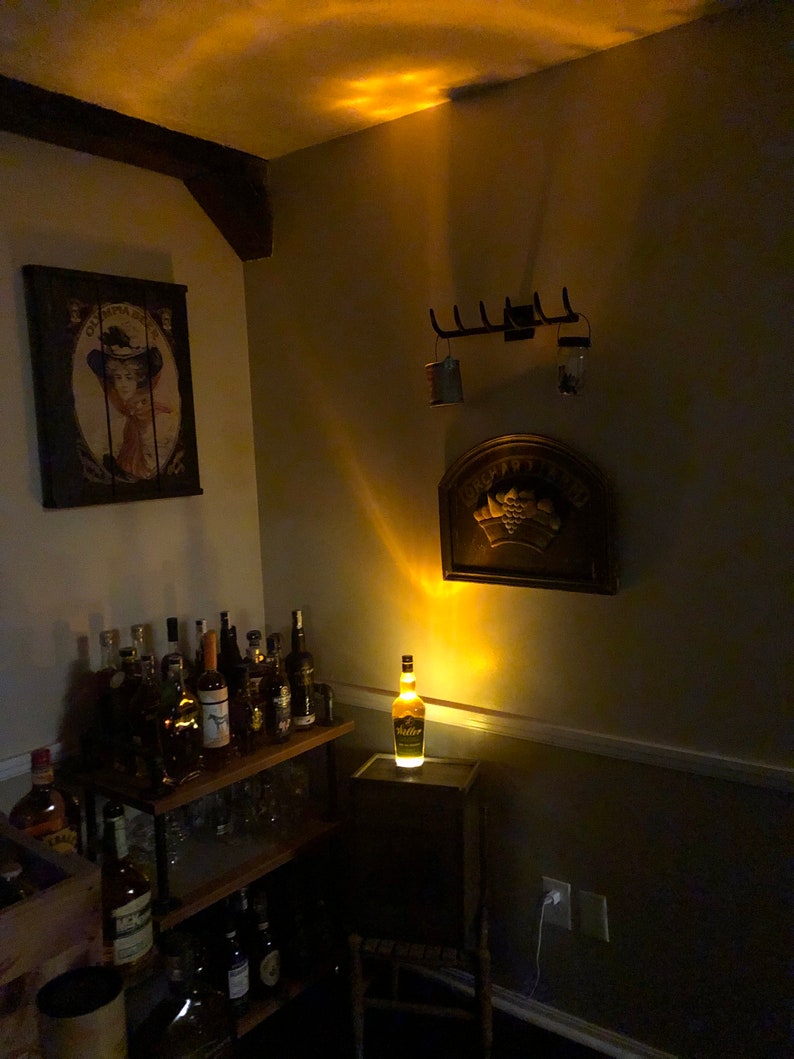 May include: A room with a drinks cabinet and a bottle of liquor lit up on a small wooden stand. A vintage painting and a decorative wall hanging are also visible. The room is dimly lit.