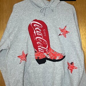 May include: Gray Gildan Unisex L hoodie with a large red Coca-Cola cowboy boot graphic. The boot is decorated with a paisley pattern and the Coca-Cola logo. Three red star accents are also on the hoodie.
