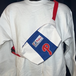 May include: White and blue fanny pack with the Philadelphia Phillies logo. The pack has a red strap and a white body with vertical stripes. The Phillies logo is a red "P" on a blue background.