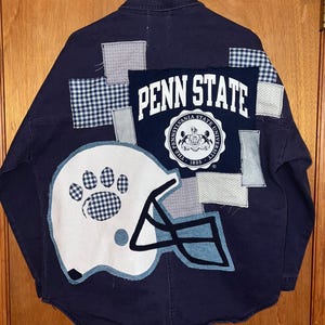 May include: Navy blue canvas jacket with patchwork details. Features a large "PENN STATE" design, a football helmet, and a paw print. The jacket is size M, measures 25" x 28", and is new with tags.