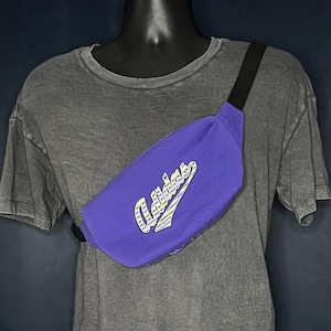 Op de afbeelding: Een paarse heuptas met het Adidas-logo in witte en gele letters. De tas heeft een zwarte band en wordt over het lichaam gedragen. De heuptas is gemaakt van duurzaam materiaal en is ontworpen voor het dragen van essentials.