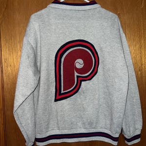 May include: A gray sweatshirt with a large maroon and navy blue "P" logo on the back. The sweatshirt has a collared neckline and navy blue and red striped trim on the cuffs and hem. The garment is hanging on a white hanger.