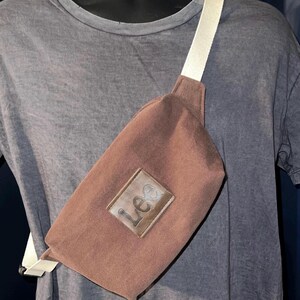 Puede incluir: Un bolso de cintura marrón con una correa blanca, usado cruzado. El bolso tiene un parche rectangular de cuero con la palabra "Lee" en relieve. El bolso de cintura está hecho de un material resistente y se usa sobre una camiseta gris.