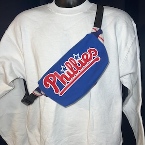 May include: A blue fanny pack with the word "Phillies" in red script with a white outline. The fanny pack has a black strap and a white and red patterned accent. The fanny pack is displayed on a white sweatshirt.