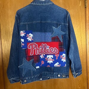 May include: A blue denim jacket with patches featuring the word "Phillies" in red and white script. The patches include star shapes and a plaid pattern. The jacket is hanging on a white hanger.