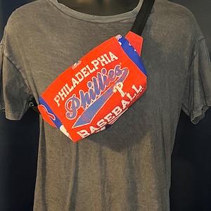 May include: Red and blue fanny pack with the text "PHILADELPHIA Phillies BASEBALL". The bag has a black adjustable strap and a zipper closure. The fanny pack is made of a durable fabric.