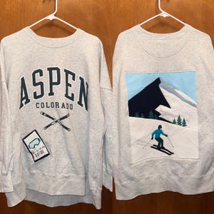 May include: A light grey sweatshirt featuring the text "ASPEN COLORADO" in dark blue. Below the text are crossed skis and a patch with ski goggles and a barcode. The back shows a skier on a snowy mountain scene.