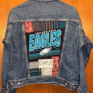 May include: A blue denim jacket with a Philadelphia Eagles logo patch on the back. The jacket is 20 inches wide and 21 inches long. The patch features the team name and logo in teal and white.