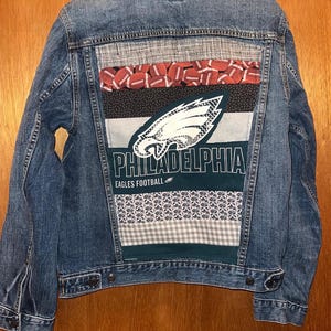 May include: A blue denim jacket with a patchwork design on the back. The patchwork includes footballs, a Philadelphia Eagles logo, and the text "PHILADELPHIA EAGLES FOOTBALL". The jacket is 20 inches wide and 24 inches long.