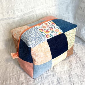 Könnte beinhalten: Ein quadratischer Patchwork-Beutel mit Reißverschluss. Der Beutel besteht aus verschiedenen Stoffen in Blau-, Rosa-, Pfirsich- und Blumenmustern. Der Reißverschluss ist leuchtend orange.