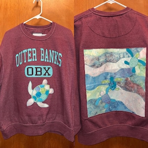 May include: Maroon sweatshirt with "Outer Banks OBX" text in teal. A turtle applique is on the front. The back features a square patch with waves and turtles. The sweatshirt is hanging on a white hanger.