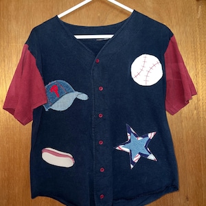 May include: A navy blue baseball jersey with red sleeves, decorated with patches of a baseball, a baseball cap, a star, and a hot dog. The jersey is buttoned up and hanging on a white hanger.