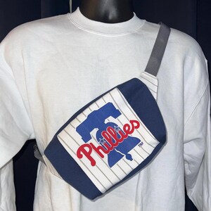 May include: A navy blue and white fanny pack with the Philadelphia Phillies logo. The bag has a gray strap and is worn across the chest. The Phillies logo is in red and blue.
