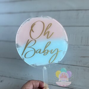 以下が含まれることがあります： ピンクとブルーのグラデーションデザインの丸い透明アクリルケーキトッパー。"Oh Baby"という文字が金色の筆記体で書かれています。