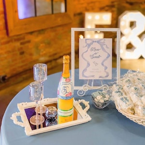 May include: A white ornate tray holding a champagne bottle with a custom label, two tall glass candle holders, and a small glass bowl. A framed sign reads Love is Sweet with a blue ribbon illustration, next to a basket of wrapped treats.