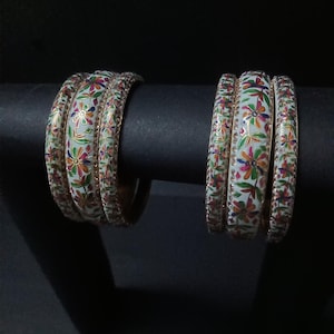 Può includere: Due set di braccialetti a fantasia floreale. Ogni braccialetto è bianco con fiori colorati e finiture dorate. I braccialetti sono esposti su un supporto nero su sfondo nero. Il design floreale comprende rosso, arancione, verde e blu.