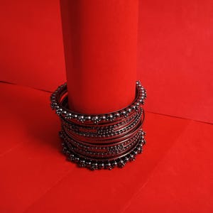 Puede incluir: Un conjunto de pulseras plateadas con detalles intrincados, incluyendo pequeñas cuentas y patrones texturizados. Las pulseras están dispuestas alrededor de un cilindro rojo, sobre un fondo rojo vibrante.