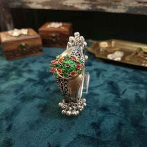 Könnte beinhalten: Ein silberfarbenes Armband mit einem verzierten zentralen Blumenmotiv mit grünen und roten Akzenten. Die Metalloberfläche zeigt komplizierte Filigranmuster und Gravuren, abgerundet durch kleine hängende silberne Perlen am unteren Rand.