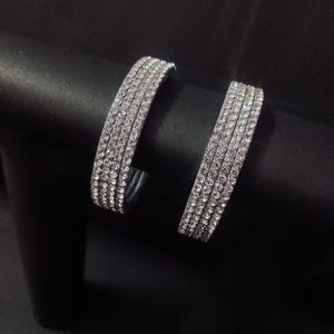 Puede incluir: Dos brazaletes plateados, cada uno incrustado con múltiples filas de pequeños pedrería brillantes. Los brazaletes se exhiben sobre un fondo oscuro y sólido, creando un contraste que resalta su brillo. La joyería parece ser nueva.