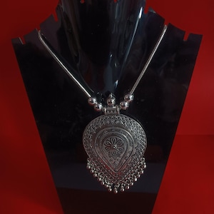 May include: Silver pendant necklace with an elaborate, teardrop-shaped pendant. The pendant has detailed embossed patterns and small dangling beads. The necklace is displayed on a black stand against a red backdrop, highlighting the jewellery's features.