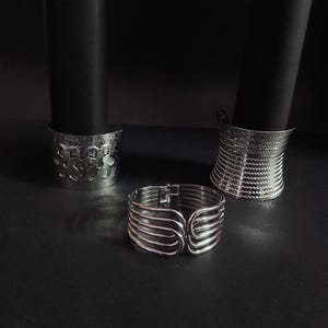 May include: Silver-toned metal bracelets with varying designs. One bracelet features a series of curved bars, while others have textured or woven patterns. The bracelets are displayed on a dark surface, with black cylindrical objects in the background.