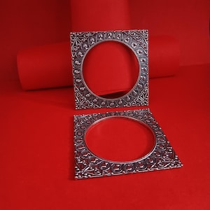 Peut inclure: Deux bracelets carrés argentés avec des motifs ornés et complexes. Chaque bracelet a une ouverture circulaire au centre. Les bracelets sont présentés sur un fond rouge vif, créant un contraste saisissant.