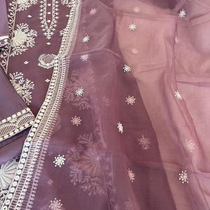 May include: A lavender-colored embroidered salwar kameez set. The tunic and pants are a solid lavender, with intricate white floral embroidery along the neckline, cuffs, and hem. A sheer, matching dupatta with scattered white floral accents completes the ensemble.